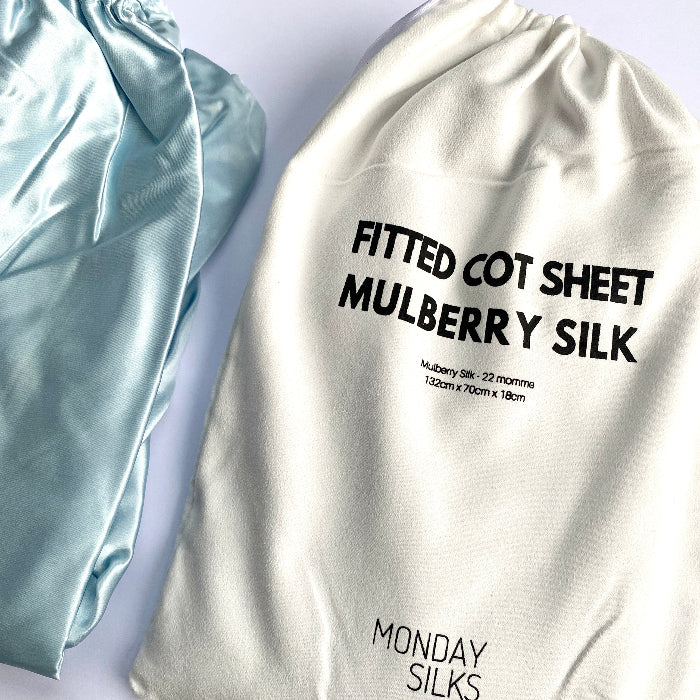 A sky blue silk cot sheet displayed next to its packaging, which is labeled 'FITTED Cot Sheet Mulberry SILK' with the brand 'Monday Silks'.