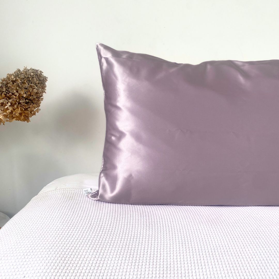 A lilac silk pillowcase on a bed with a white sheet, the pillowcase has a subtle sheen and a zipper visible on the side.