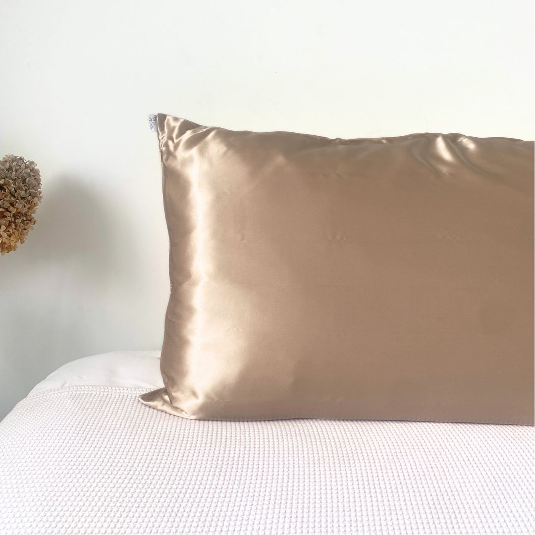 A champange colored silk pillowcase on a bed, with a white checkered sheet visible beneath it. A dried plant decoration is partially visible in the top left corner.