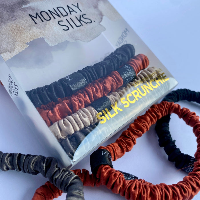 Packaging of Monday Silks skinny silk scrunchies in four different colors: black, brown, gray, and bronze, displayed on a white background.